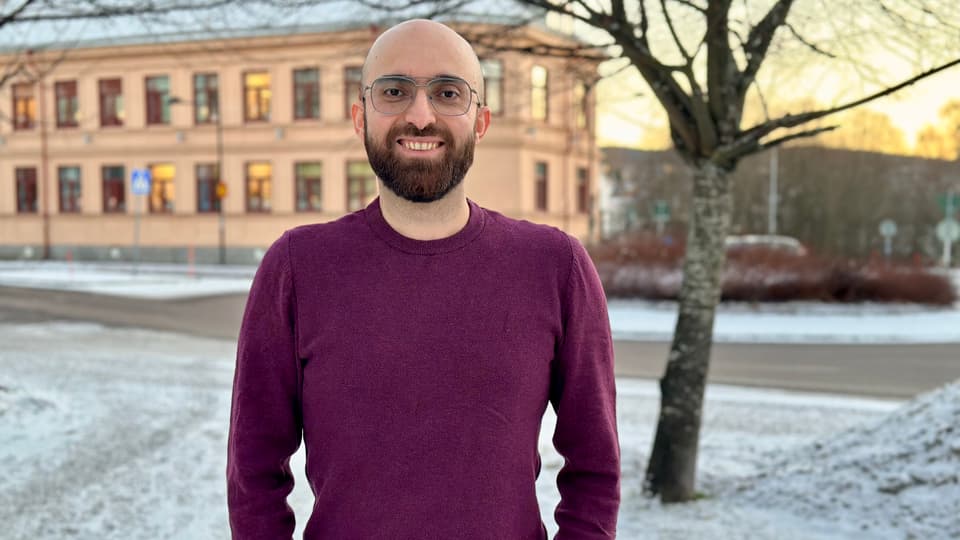 Leende man med rakat huvud, brunt skägg och glasögon. Han har vinröd tröja och står utomhus. Årstiden är vinter.