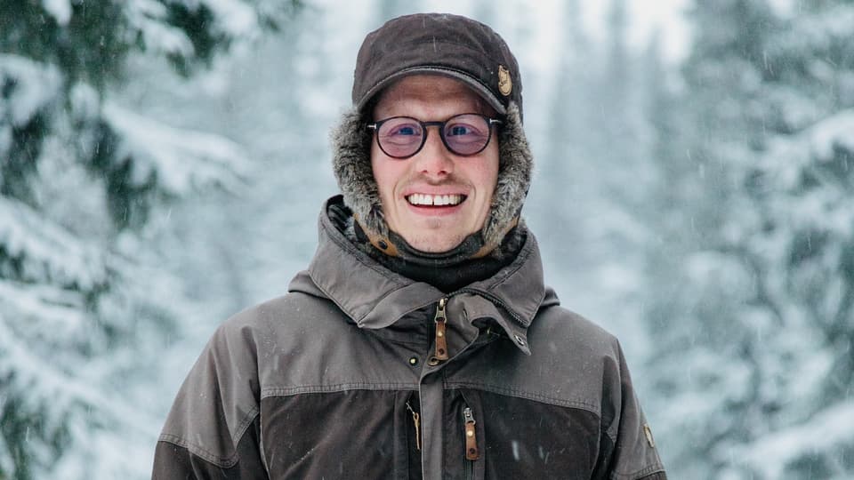 Glad ung man i tjocka vinterkläder står utomhus i vinterlandskap med snöklädda granar i bakgrunden
