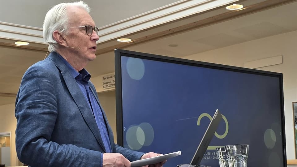 Gråhårig man i kavaj håller föredrag framför en bildskärm