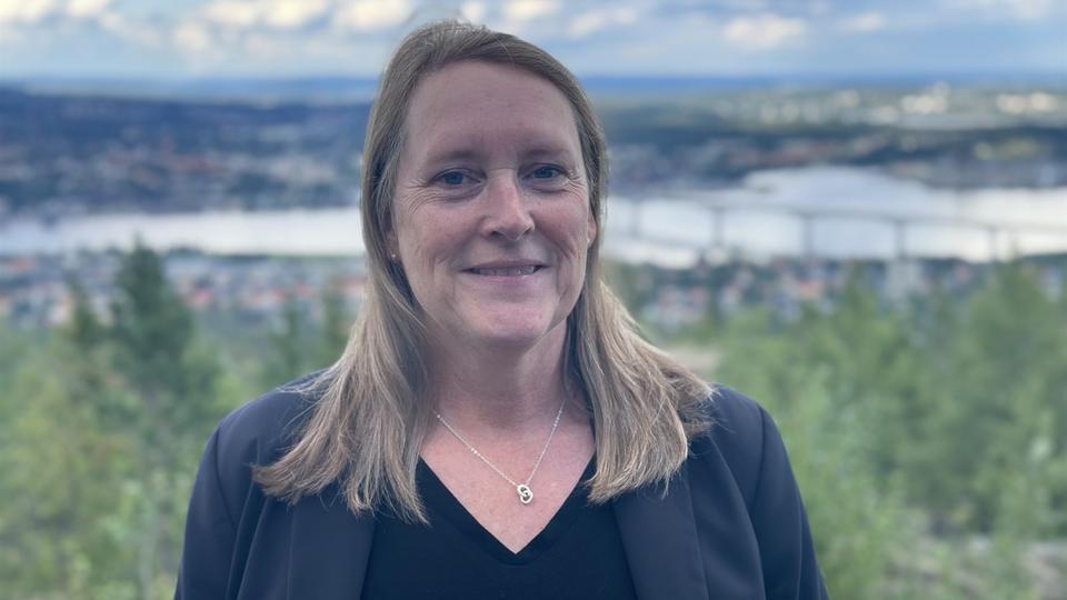En kvinna med blont axellångt hår, svart tröja och mörkblå kavaj med en naturskön vy med skog, hav och en bro i bakgrunden.