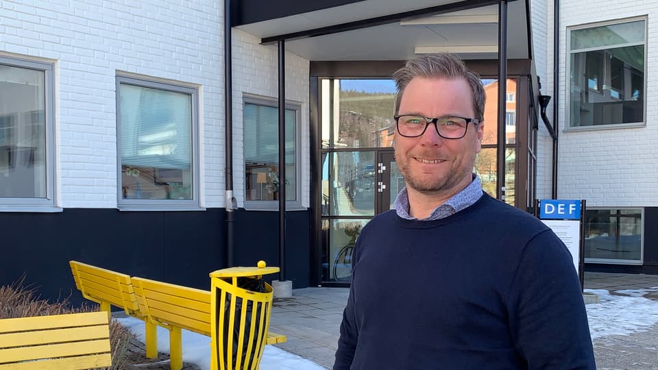 En man i glasögon och marinblå tröja som står utanför ett vitt hus med gula bänkar i bakgrunden