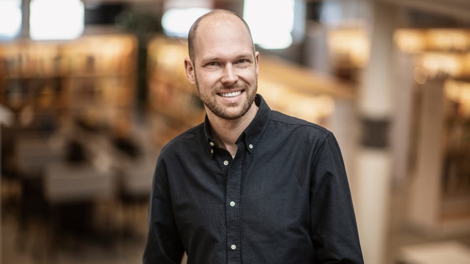 En man i svart skjorta med vita knappar ler mot kameran