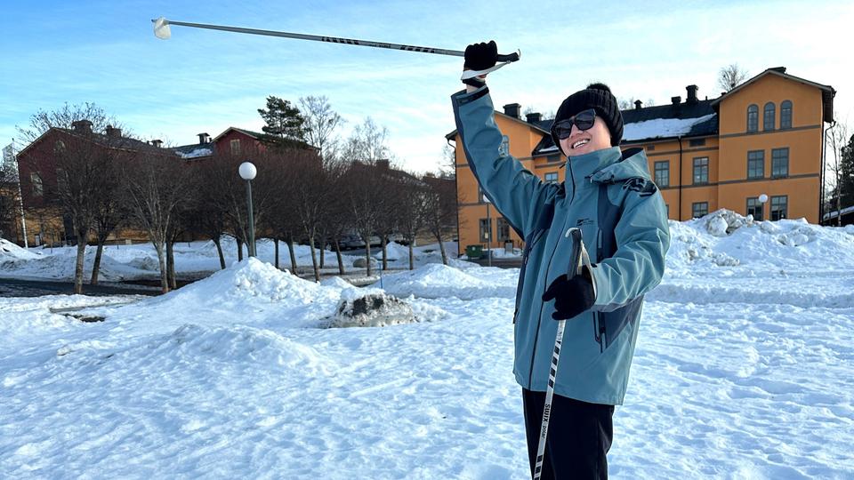 Young man with poles in his hands stands on a snow-covered field and raises one hand in the air and smiles