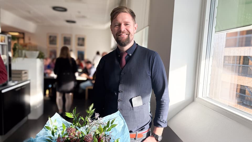 En man med kort brunt hår ler med blommor framför sig