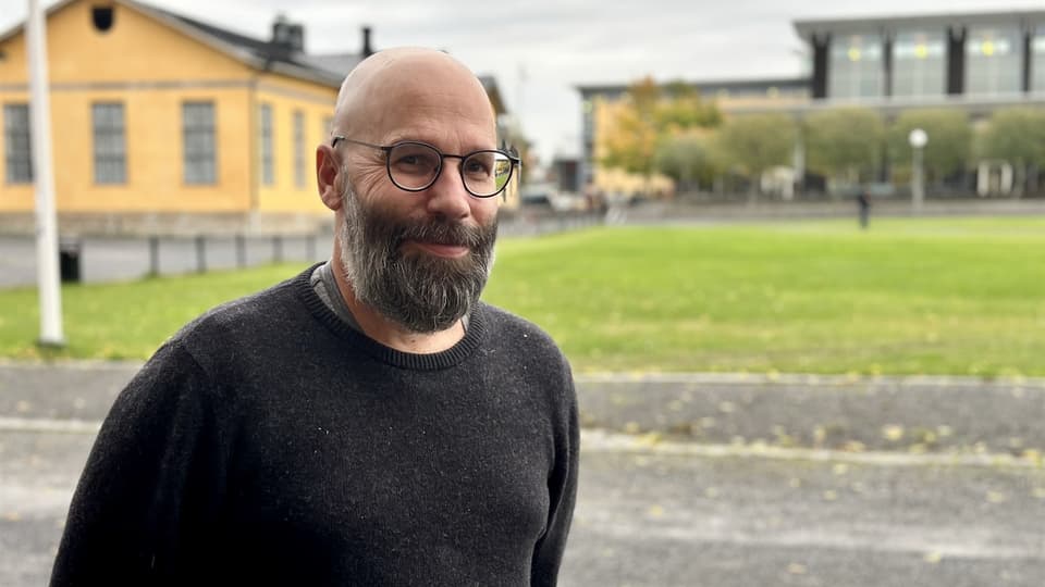 En man med glasögon och en mörkgrå tröja som står utanför framför gula byggnader och ler in i kameran