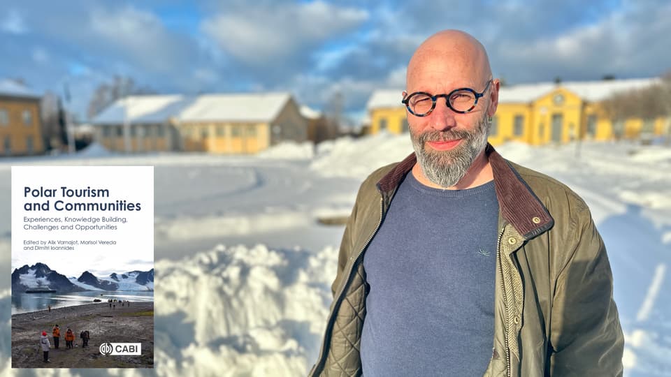 En man i glasögon, blå tröja och grön jacka står utomhus i ett snigt landskap med gula byggnader i bakgrunden. En bild på ett bokomslag är inklippt i bilden. 