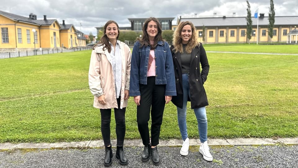 Tre unga kvinnor i ytterkläder står framför en stor gräsyta med hus i bakgrunden. De ler in i kameran. 