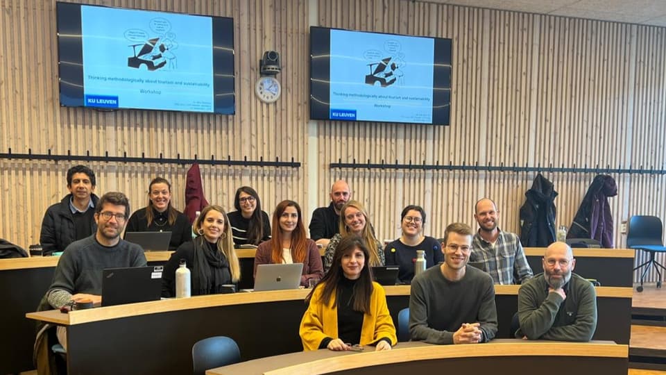 A dozen people sit in a lecture hall and look into the camera with their computers in front of them and two monitors in the background. 