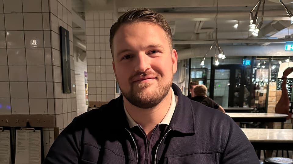 Man med mörkt kort hår, skägg och mörkblå kofta tittar in i kameran.