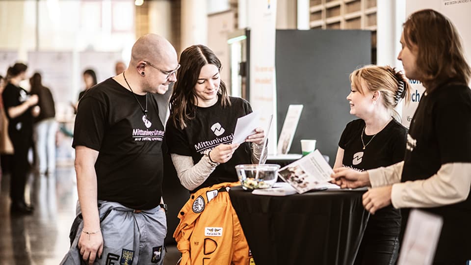 Två manliga och två kvinnliga studenter i svarta Mittuniversitetet t-shirts står runt ett bord och läser på ett vykort. 