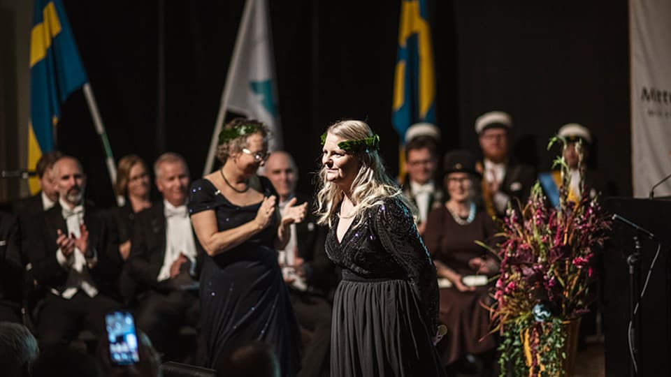 En finklädd kvinna med lagerkrans står på en scen. 