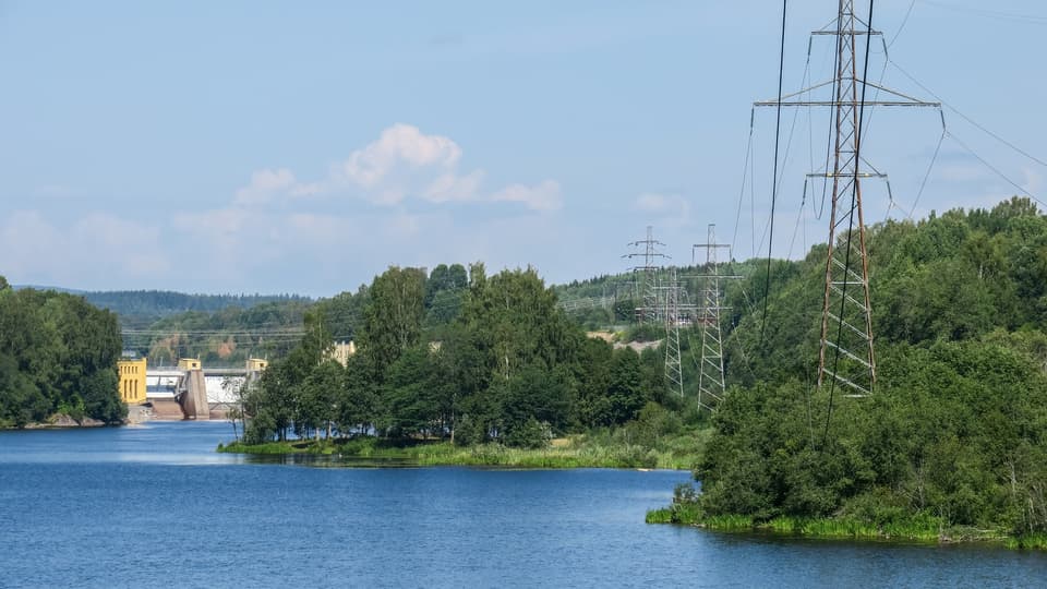 Elnätsstolpar står i ett landskap med skog och ett vattendrag där ett vattenkraftverk syns i bakgrunden.