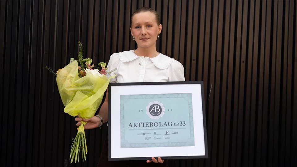 En kvinna tittar in i kameran. Hon håller i en blombukett och i en tavla.