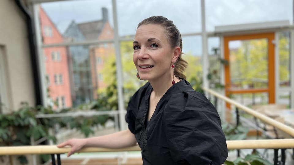 En kvinna tittar in i kameran och ler. I bakgrunden syns en glasvägg med husfasader utanför. 