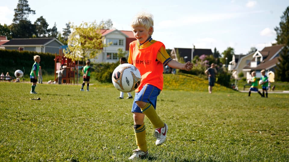En pojke med blondt hår i fotbollskläder jonglerar med en fotboll på en gräsplan med andra barn och villor i bakgrunden. 