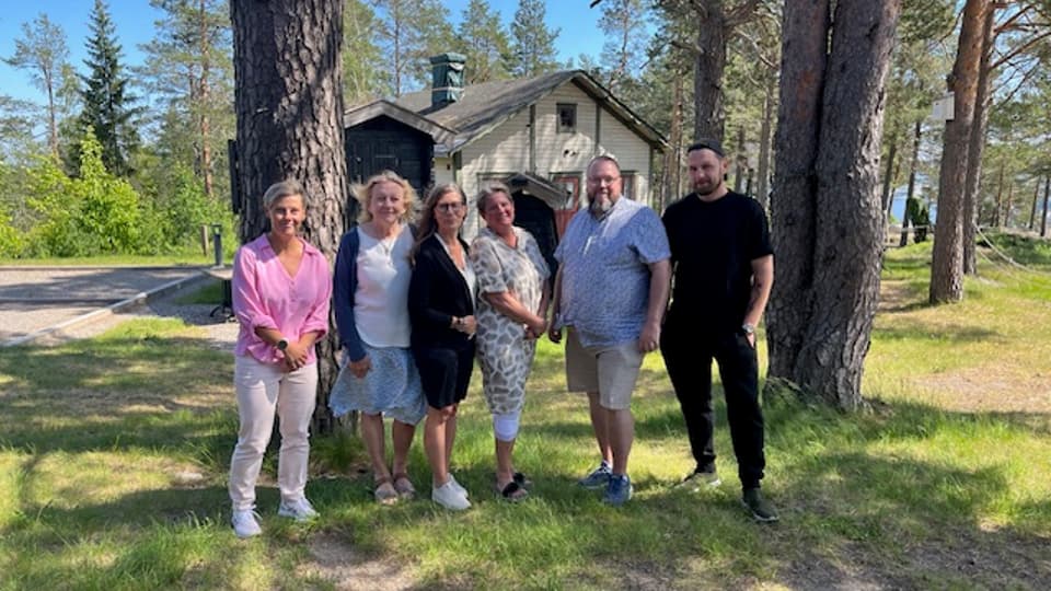 Fem personer blir fotograferade utomhus framför två stora träd och en grön gräsmatta