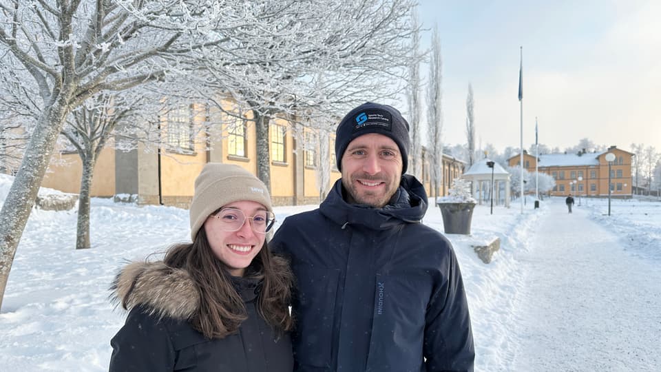 Två personer står framför frostiga träd och byggnader klädda i vinterkläder.