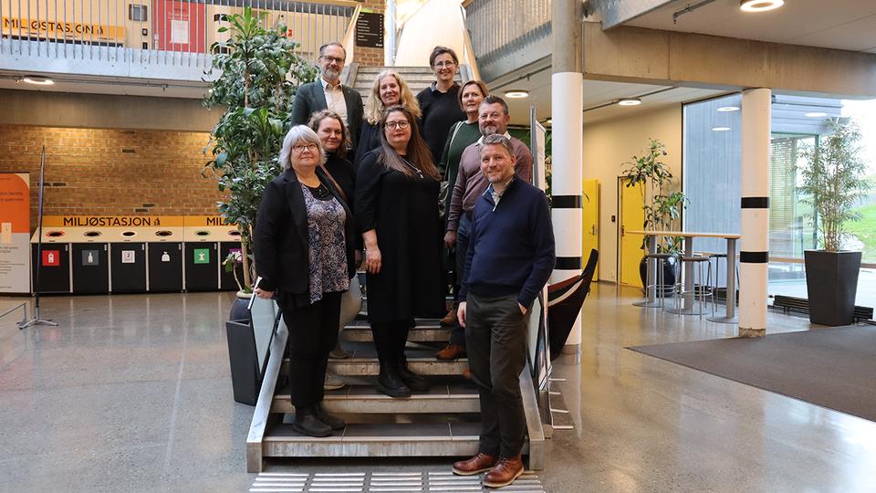 Nio personer står i inomhusmiljö i en trappa på de nedersta trappstegen. Runt omkring dem finns öppna ytor.