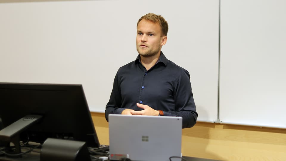 en man i mörkblå skjorta står framför en vit vägg. 