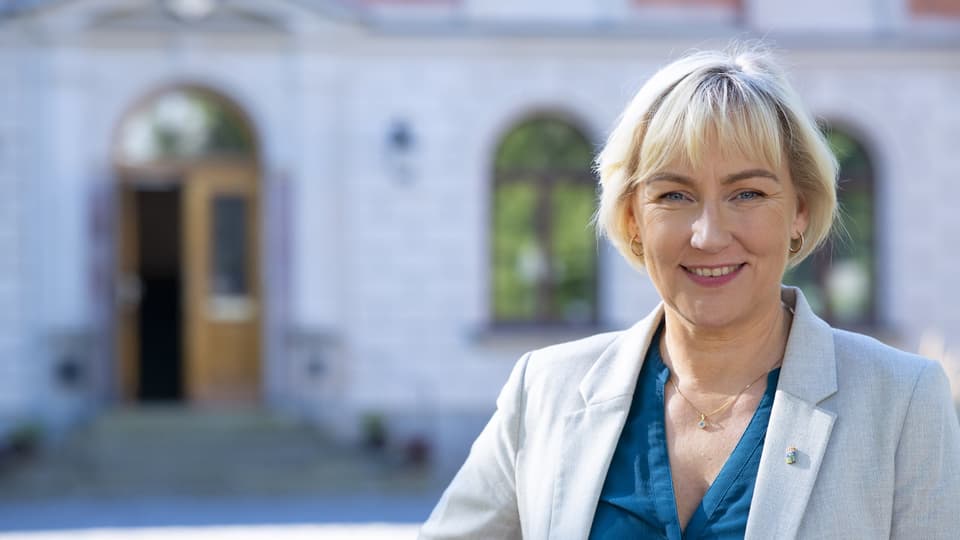En kvinna i grå kavaj och blå blus står framför en byggnad.