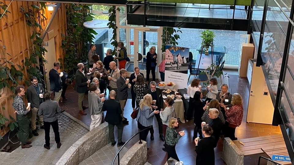 Ett 30-tal människor står i smågrupper och pratar. Många har smörgås eller frukt i handen. Bilden är tagen snett uppifrån och i förgrunden syns ett metallräcke.
