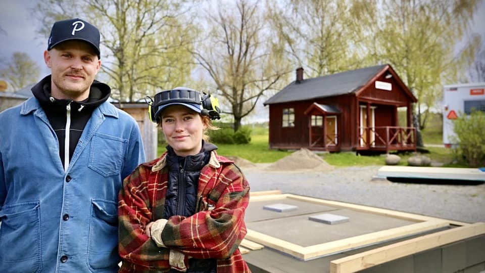 Två personer, en man och en kvinna, står utomhus med två små husgrunder bakom sig. 