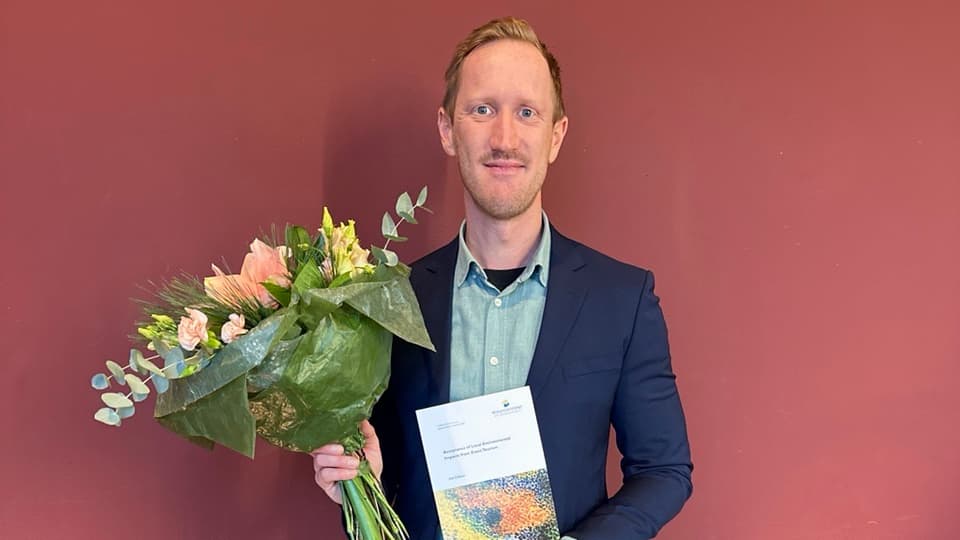 Man iklädd blå kostym och grön skjorta står framför en röd vägg och håller i en blombukett och en avhandling. 