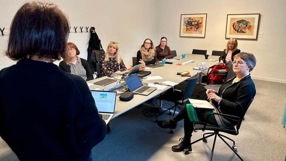 Sex kvinnor sitter runt ett konferensbord och tittar mot en annan kvinna som står upp med ryggen mot kameran.
