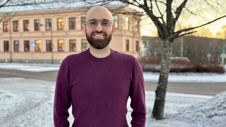 Leende man med rakat huvud, brunt skägg och glasögon. Han har vinröd tröja och står utomhus. Årstiden är vinter.