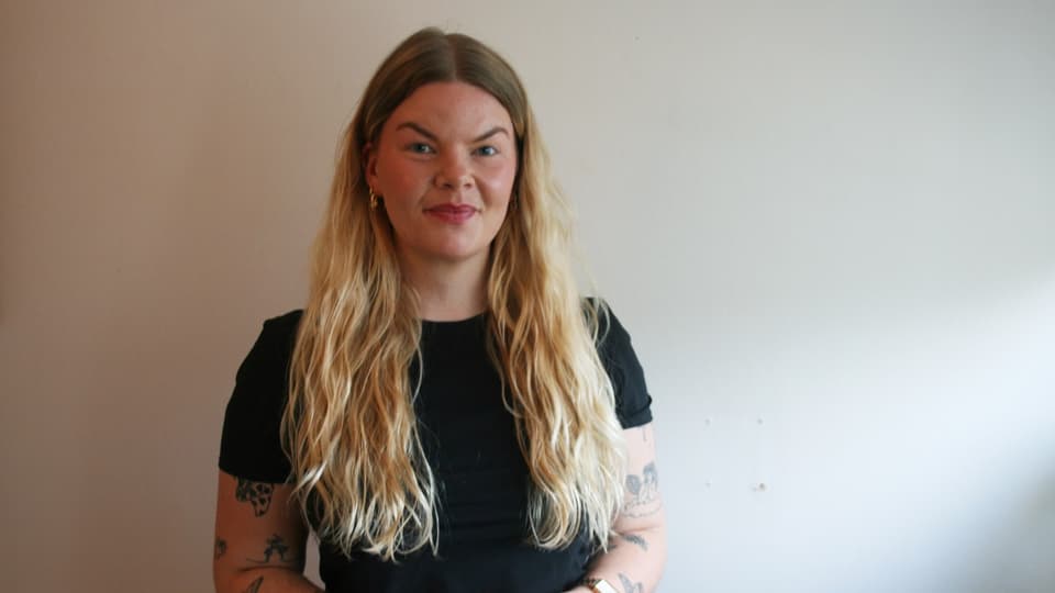 Kvinna i svart kortärmad tröja och långt blont hår står framför en beige vägg. 