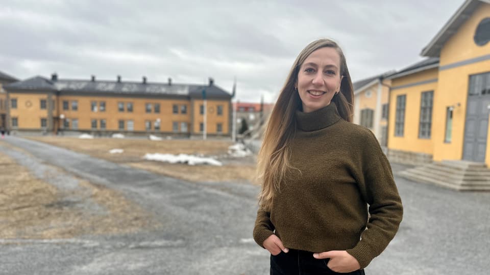 Kvinna i brun polotröja och svarta byxor står utomhus framför gula stora byggnader