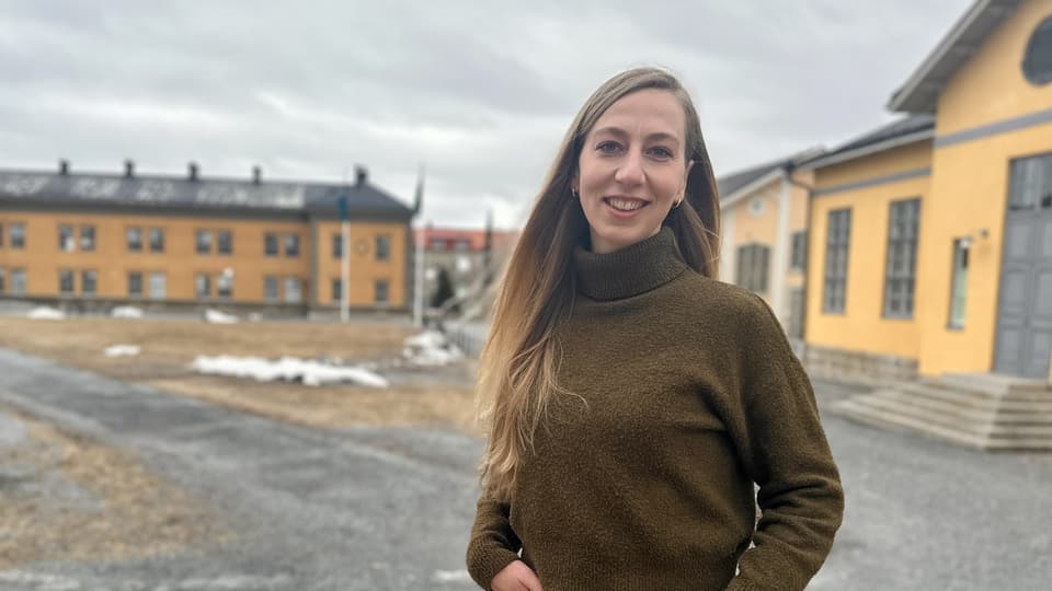 Kvinna i brun polotröja och svarta byxor står utomhus framför gula stora byggnader