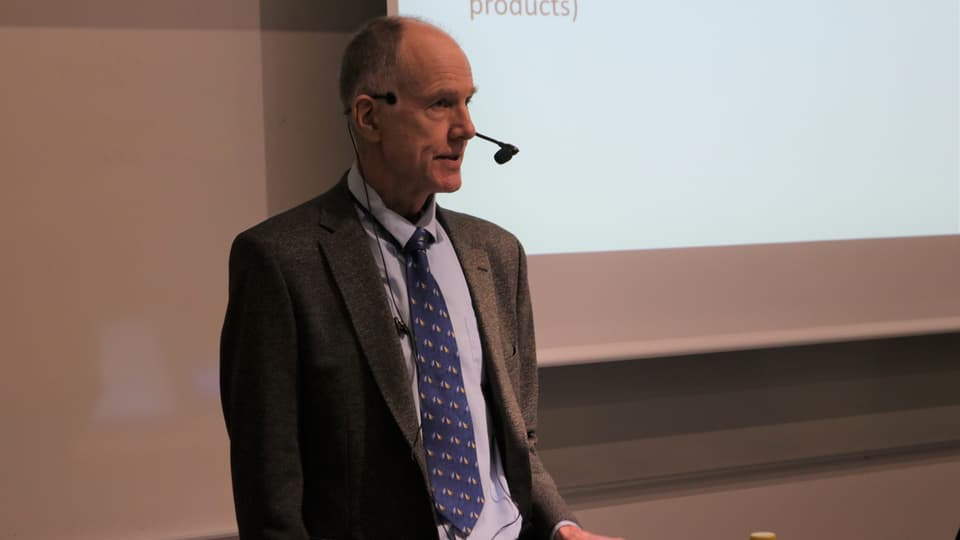 En man i kavaj och slips, med en mic som står framför en whiteboard