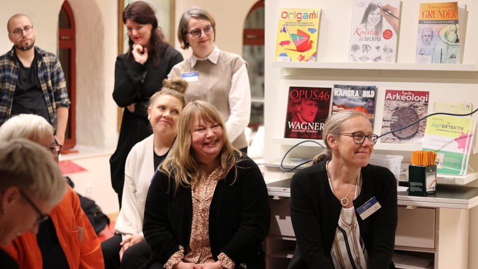 Åtta personer i biblioteksmiljö, några sitter och några står upp, och bakom dem finns vägghyllor med böcker. Vissa av personerna har namnskyltar på sig.   