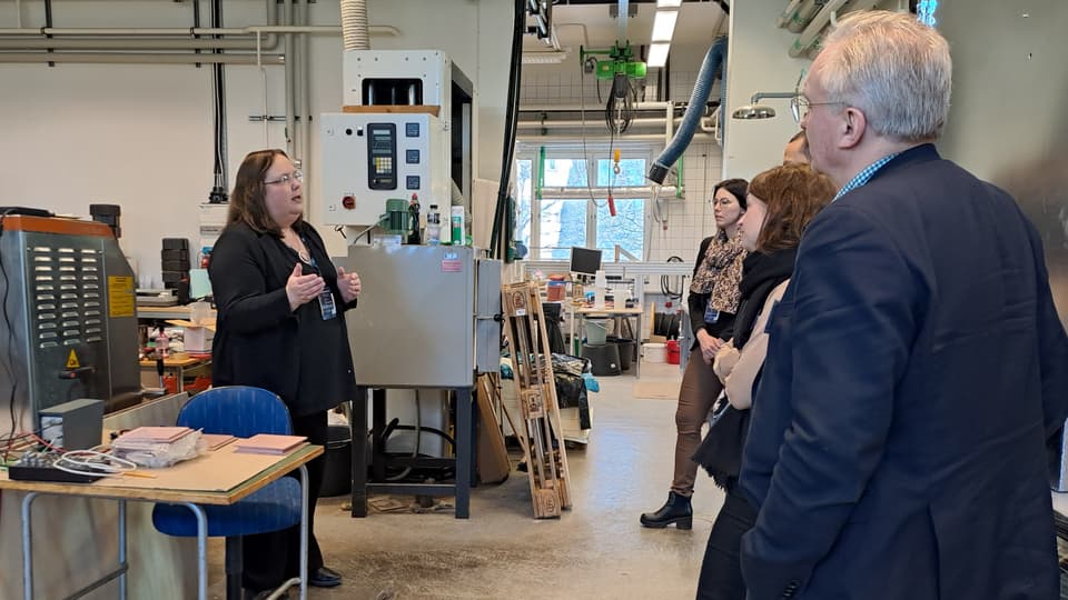 En grupp personer i en verkstad lyssnar på en kvinna som talar.