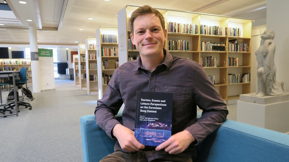 Ung man sittandes i ett bibliotek hållandes en bok. Han har en kort frisyr och bär grå skjorta. 