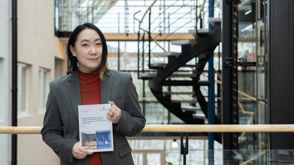 En kvinna inomhus håller i en avhandling med båda händer och ler mot kameran
