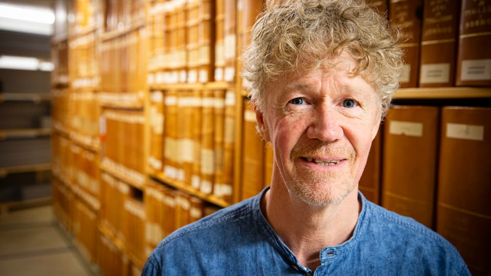 En man med blå skjorta tittar in i kameran. Bakom honom syns hyllor fyllda med gamla böcker. 