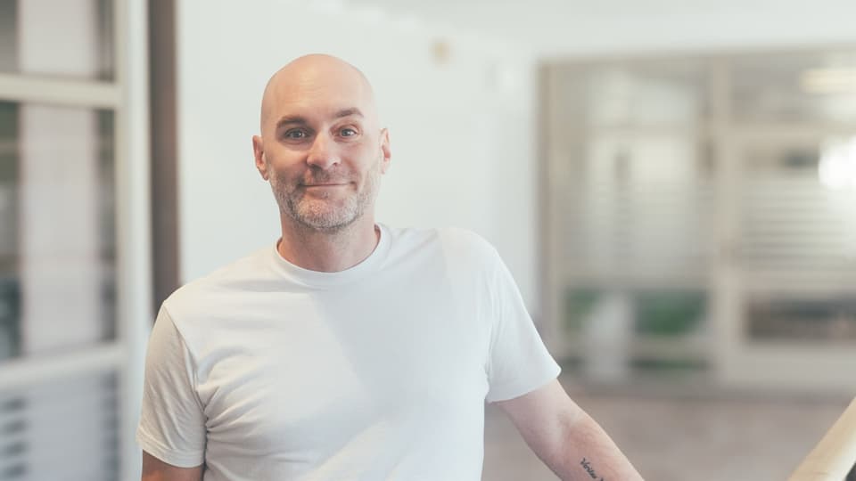 En man i en vit t-shirt står i en ljus lokal. Han ler och tittar in i kameran.,