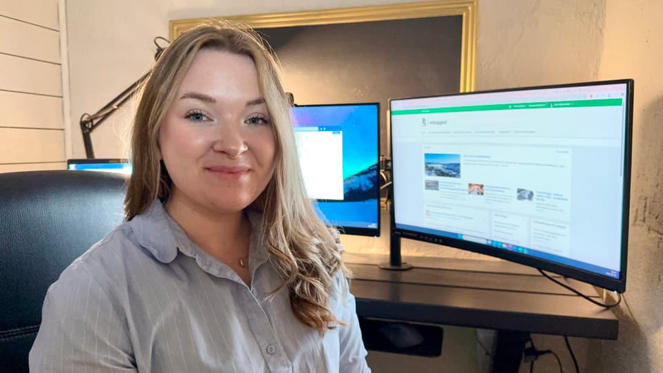 En kvinna tittar in i kameran och ler. I bakgrunden står två dataskärmar.