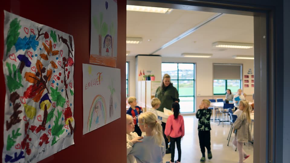 En vy av ett klassrum genom en öppen dörr. Inne i klassrummet syns en kvinnlig lärare och omkring tio barn. I förgrunden till vänster syns en röd vägg med tre teckningar
