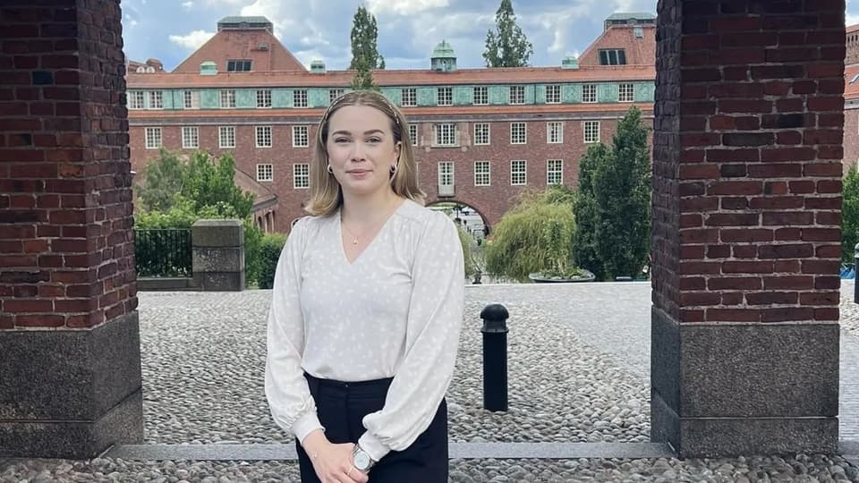 En kvinna med vit tröja och svarta byxor framför en byggnad