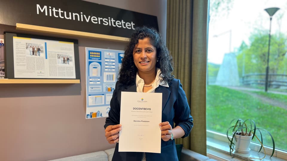 En kvinna tittar in i kameran och ler. Hon håller ett dokument i handen.