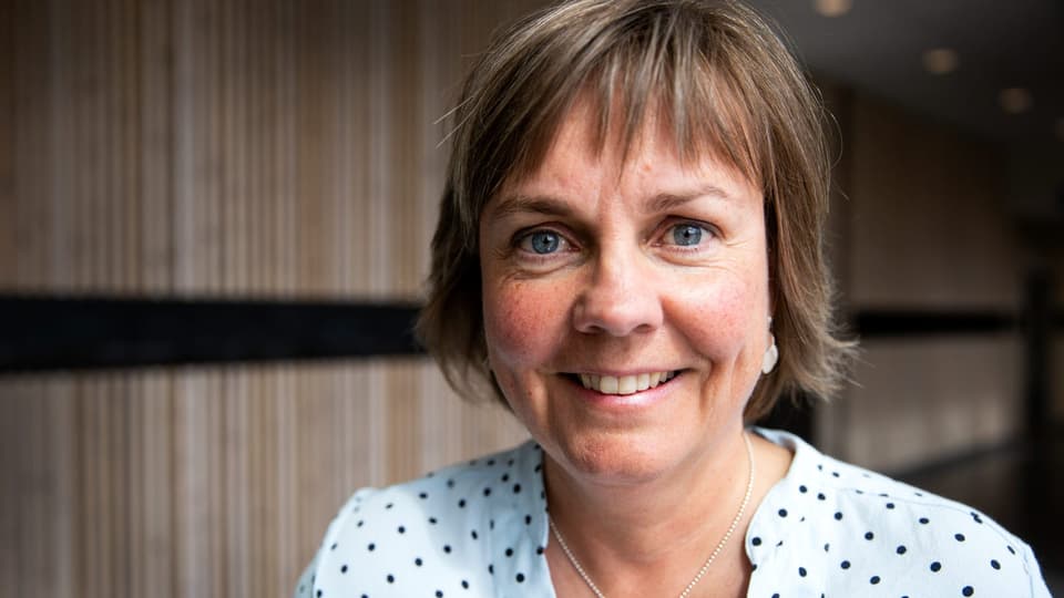 Kvinna med kortklippt frisyr och prickig blus står framför en vägg med träpanel och ler.  