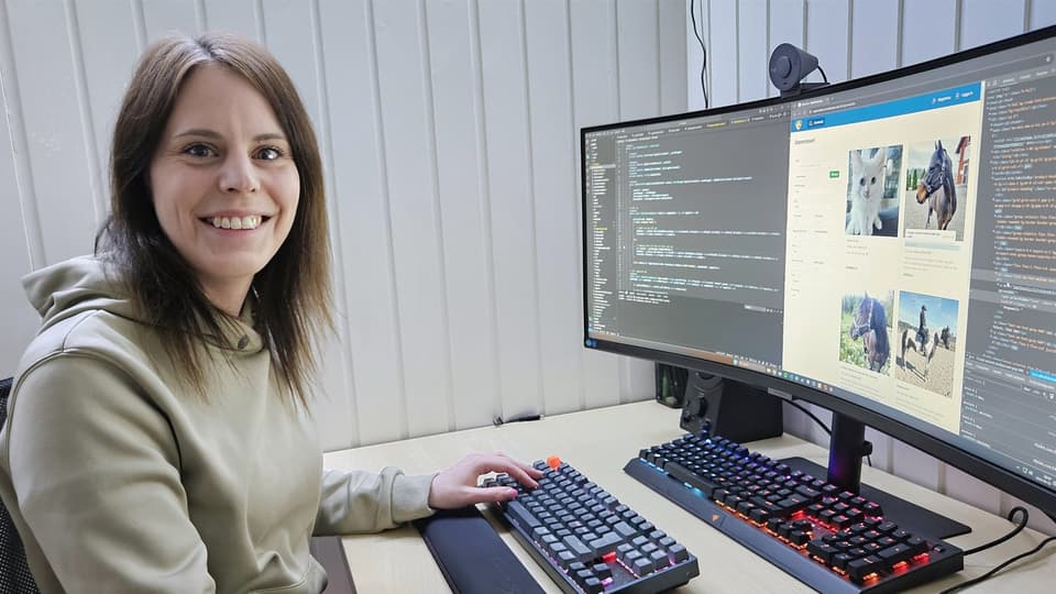 En kvinna i grå tröja sitter vid en arbetsplats med två bildskärmar framför sig. Hon ler och tittar in i kameran.