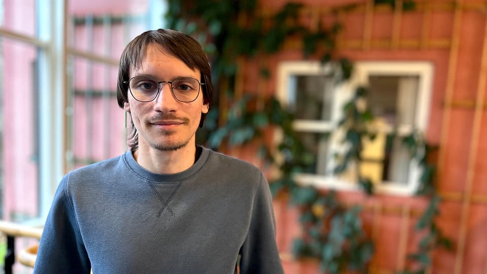 En man med brunt hår, glasögon och grå tröja står framför en rödbrun vägg med klättrande gröna växter. 