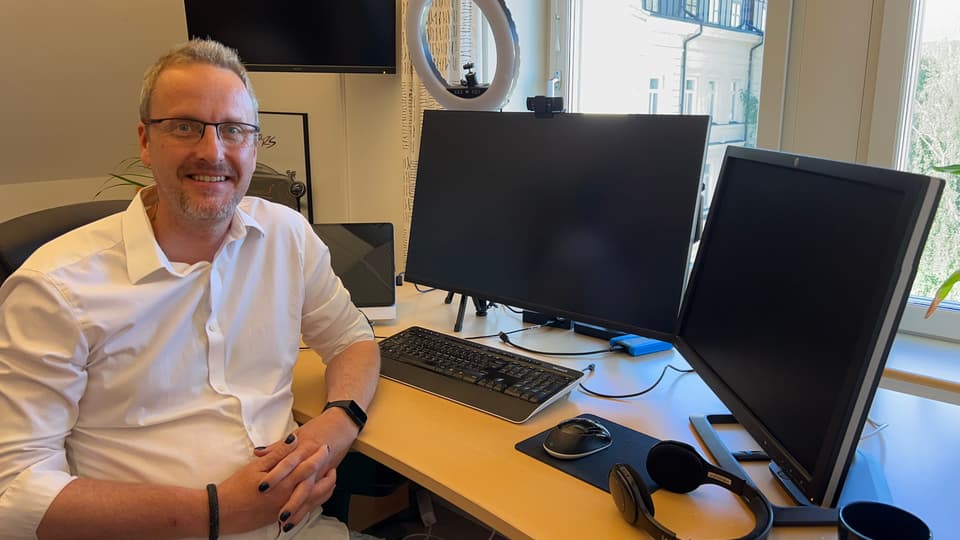 En man i glasögon och klädd i vit skjorta sitter framför några dataskärmar