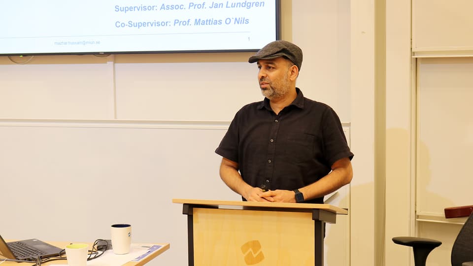 Man i svart skjorta och grå keps står och håller en presentation.