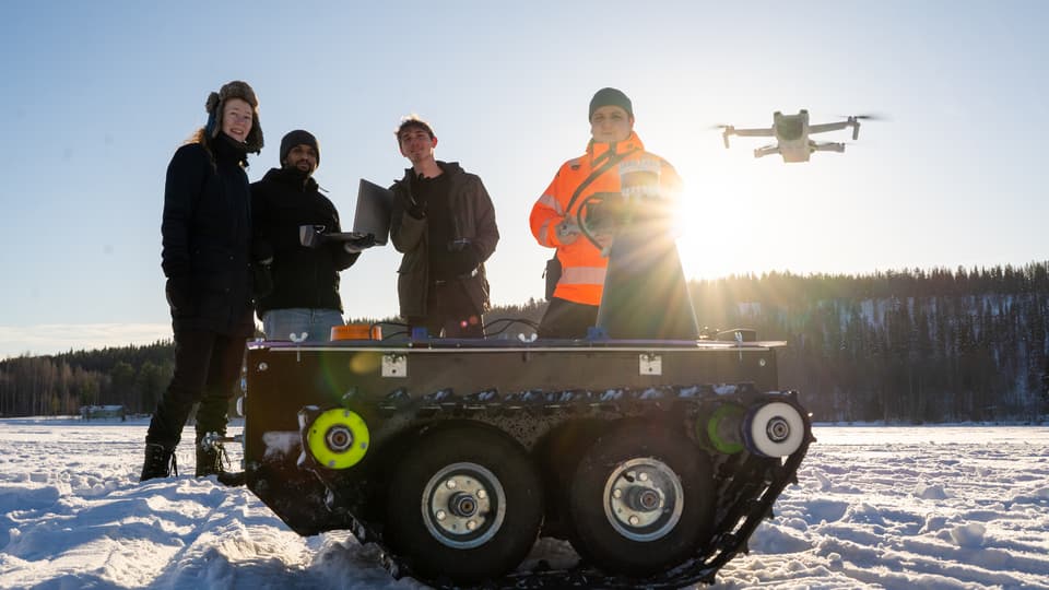  Four people are standing in a snowy landscape. A drone flies behind them and a small all-terrain vehicle stands in front of them.