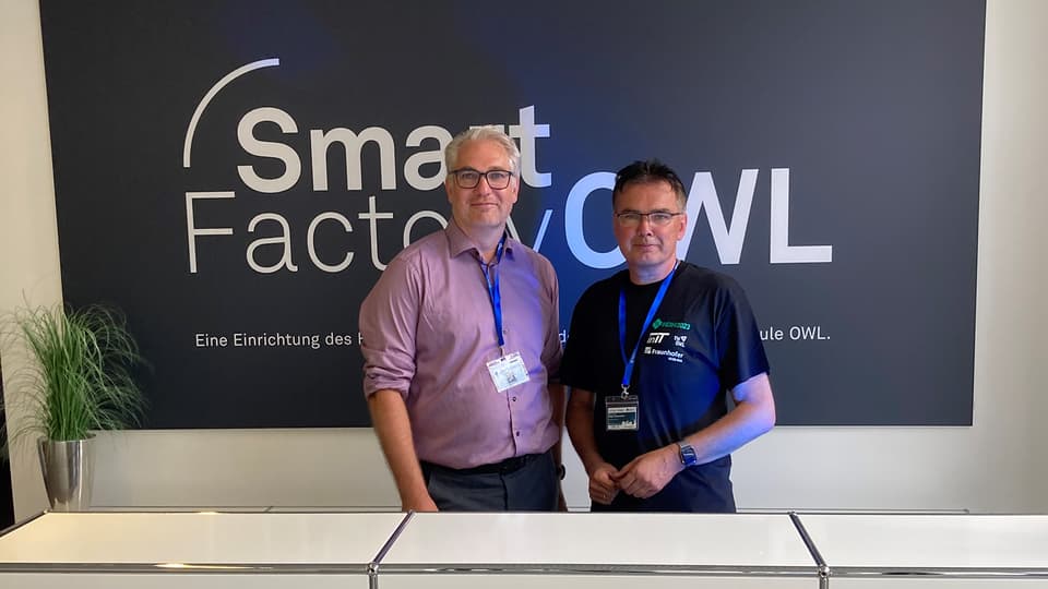 Två män står och ler mot kameran med en skylt bakom där det står Smart Factory OWL 